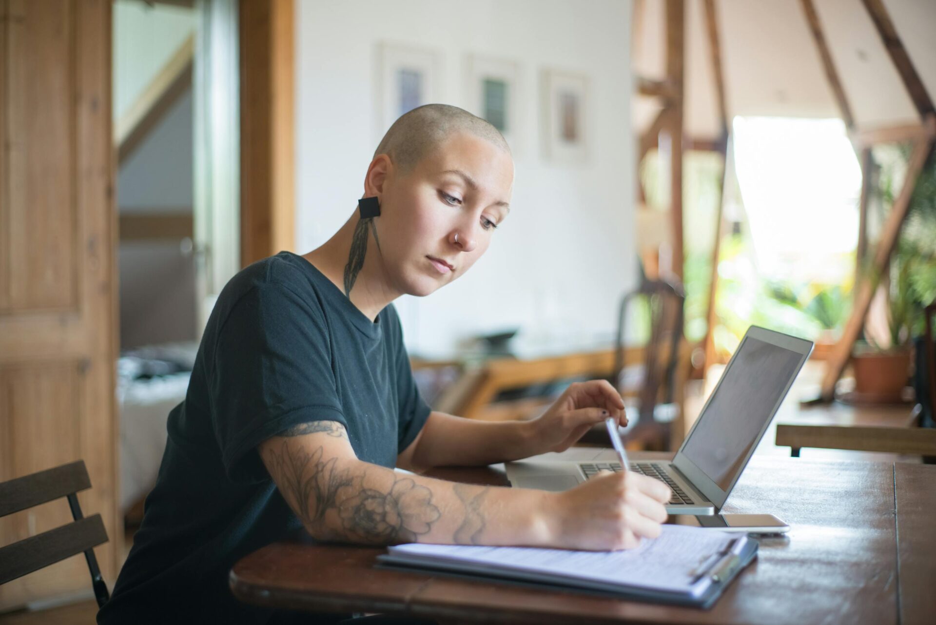 Bald woman with tattoos working on a laptop and notepad indoors. Feeling empowered after being derailed in their tech career